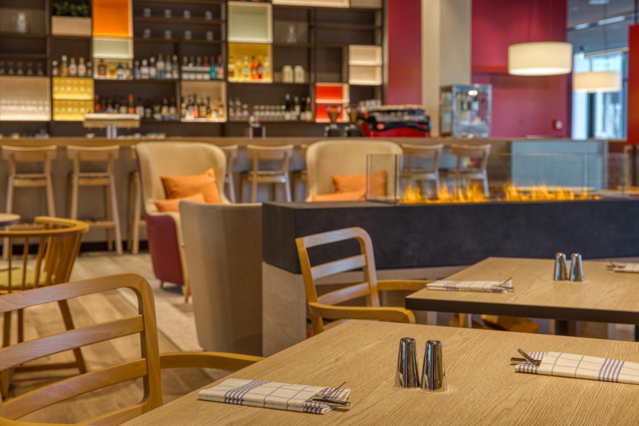 Intercityhotel Berlin Airport Ber Terminal 1 & 2 Schönefeld Eksteriør bilde The photo shows a modern restaurant or bar interior. In the foreground, there are wooden tables set with napkins and condiments. The seating includes wooden chairs and plush seating in various colors in the background. A stylish bar area is visible w